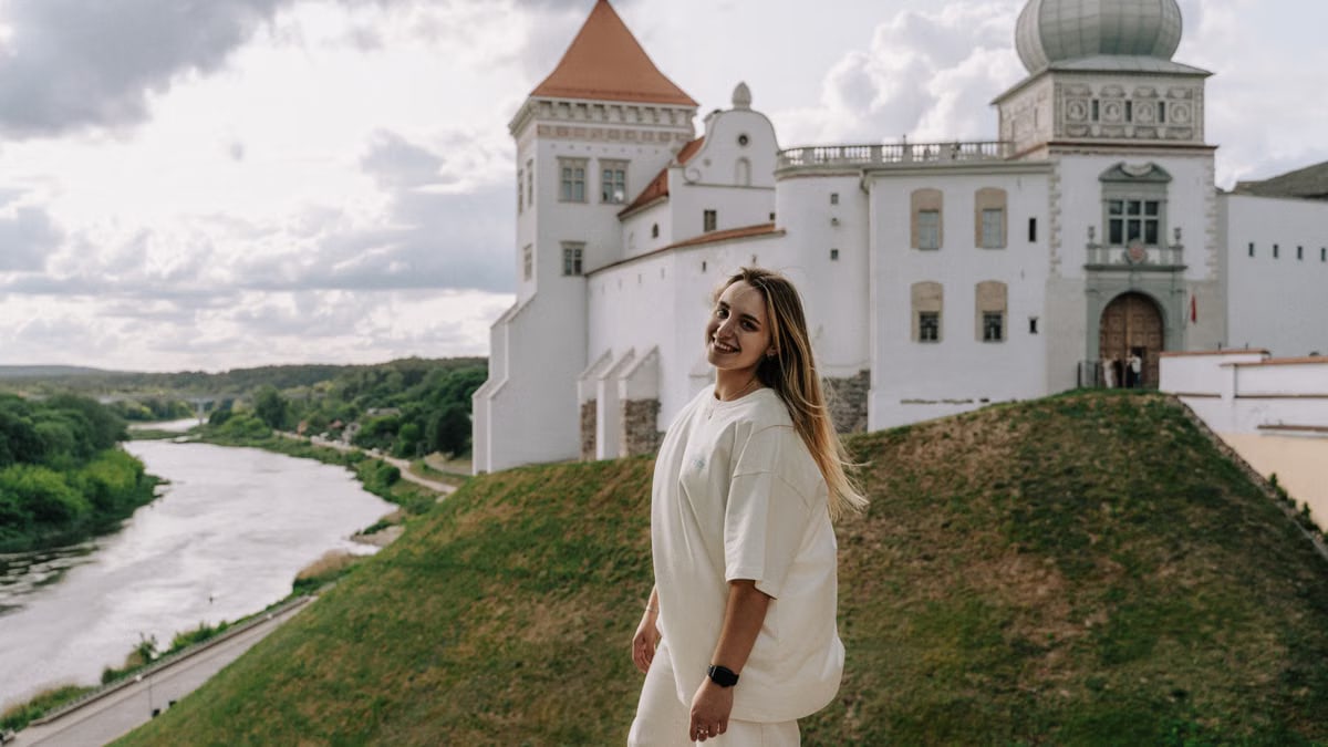 A woman enjoys a sunny day by a historic castle, with scenic river view.